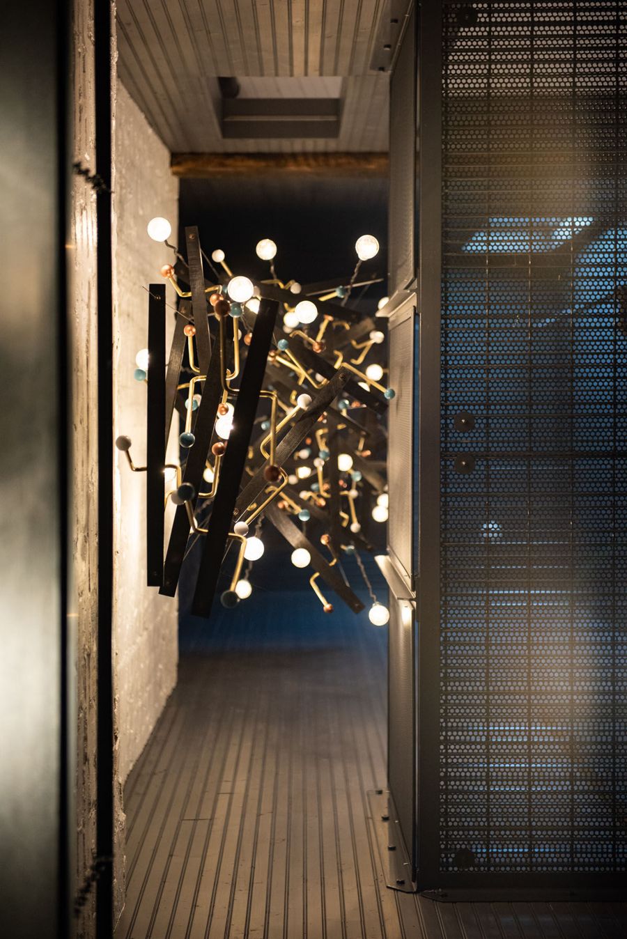 A suspended sculpture of brass branches and glowing bulbs in a dark corridor with steel mesh walls
