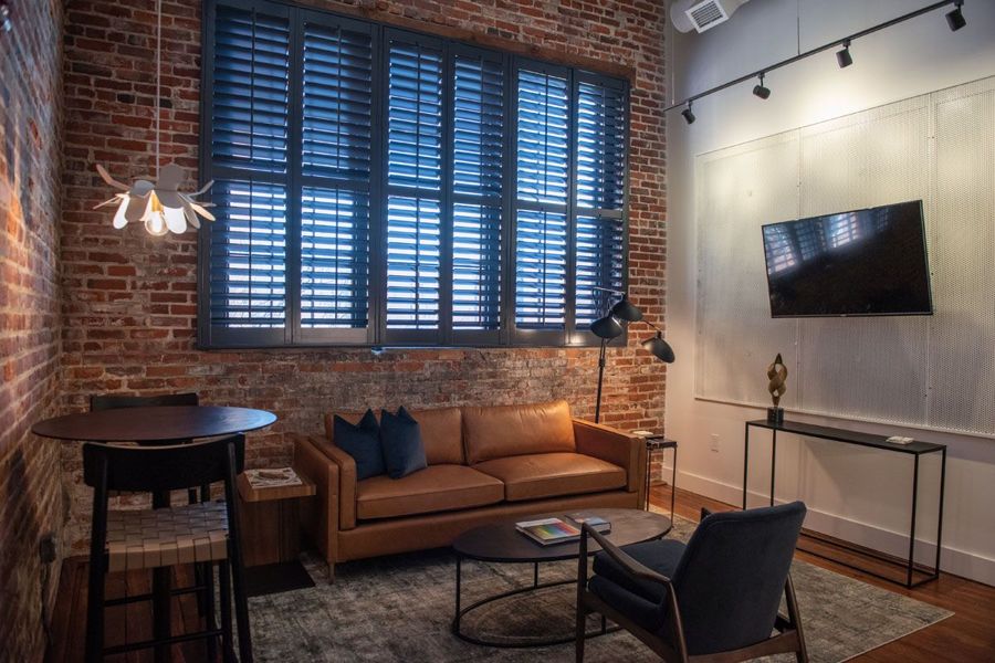Living room with a cognac leather sofa, navy armchair and bistro table beneath tall shuttered windows set in exposed brick
