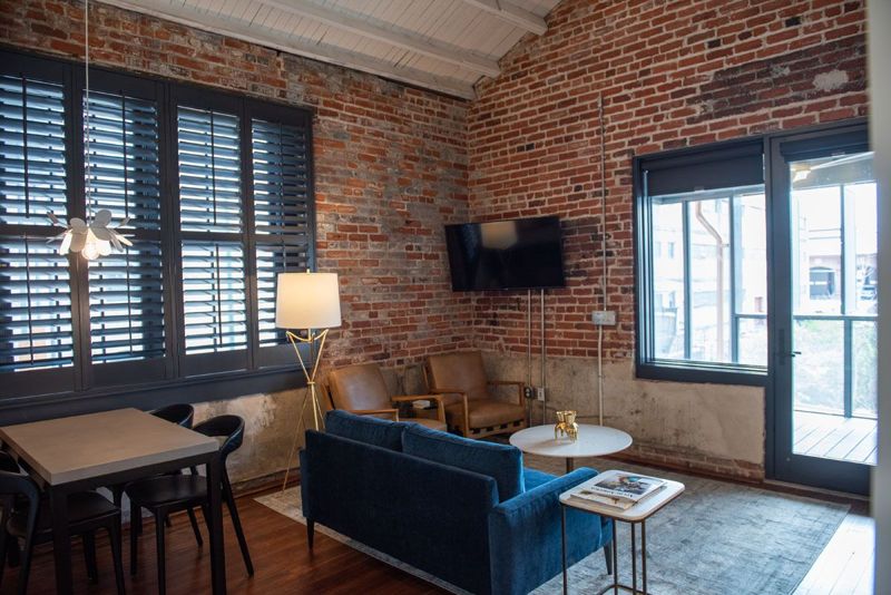 Living room with a blue velvet sofa, leather armchairs and a dining table beneath shuttered windows in exposed brick, with a door to the rear porch
